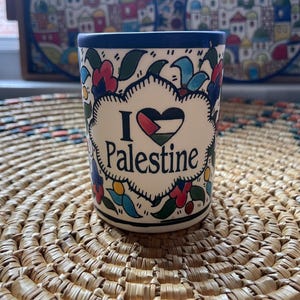 May include: A white ceramic mug with a blue rim and floral designs. The mug has a heart with the Palestinian flag and the text "I love Palestine".