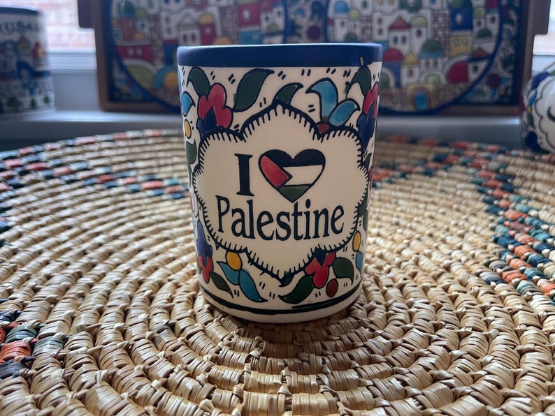 May include: A white ceramic mug with a blue rim and floral designs. The mug has a heart with the Palestinian flag and the text "I love Palestine".