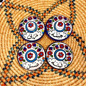 May include: Four round ceramic lids with floral designs in red, blue, and yellow. Each lid has a different Arabic word written in black on a white background. The lids are sitting on a woven straw mat.