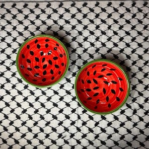 Pode incluir: Duas tigelas redondas de cerâmica com um design de melancia. As tigelas têm um interior vermelho com formas de sementes pretas e uma borda verde. Elas estão sobre um tecido estampado em preto e branco.
