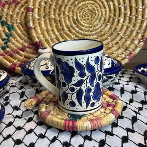 May include: A blue and white ceramic mug with a floral design sits on a woven coaster. The mug has a blue rim and a blue floral pattern with white accents. The coaster is made of natural materials and has a pink and brown color scheme.
