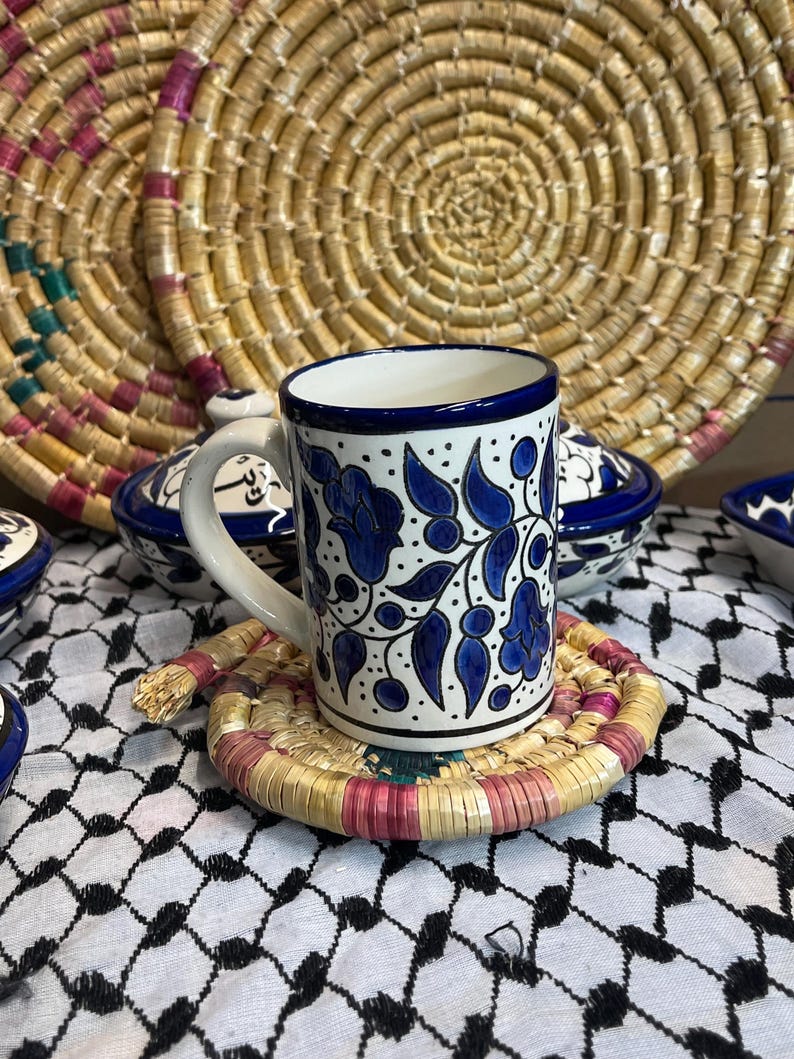 May include: A blue and white ceramic mug with a floral design sits on a woven coaster. The mug has a blue rim and a blue floral pattern with white accents. The coaster is made of natural materials and has a pink and brown color scheme.