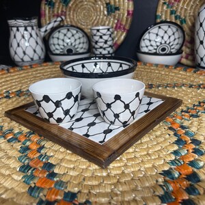 May include: A collection of white ceramic tableware with a black geometric pattern. The set includes cups, bowls, and a tray, all displayed on a woven mat. Additional decorative pieces are visible in the background.