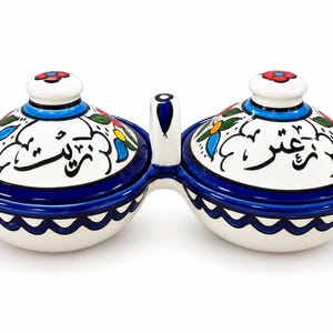May include: A double-bowl ceramic condiment server with two matching lids, featuring a white base with hand-painted blue, green, red, and yellow floral motifs and black Arabic calligraphy. The bowls are connected by a central vertical handle.