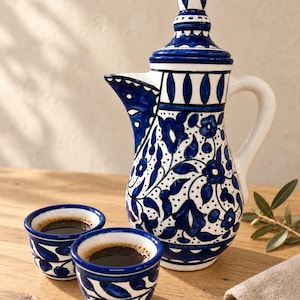 May include: A ceramic coffee pot with a pointed lid and two matching small cups, all featuring a white base with hand-painted dark blue floral and leaf patterns. The coffee pot has a curved handle and a spout, and both cups are filled with dark coffee.