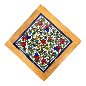 May include: Square wooden tile with a floral design. The center tile features a white background with red, blue, and yellow flowers, green leaves, and vines. The tile is framed by a blue border and a light brown wooden frame.