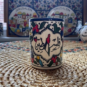 May include: A white ceramic mug with blue trim and colorful floral designs. The mug features Arabic calligraphy that reads "Wa Salam" in a heart shape.