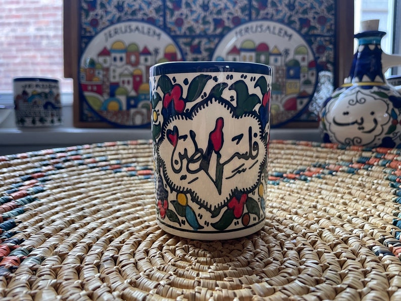 May include: A white ceramic mug with blue trim and colorful floral designs. The mug features Arabic calligraphy that reads "Wa Salam" in a heart shape.