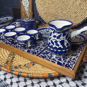May include: A wooden serving tray with a blue and white floral pattern. The tray is filled with small blue and white ceramic cups and a matching pitcher. The tray is sitting on a woven straw mat.