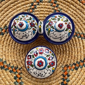 May include: Two blue and white ceramic bowls with floral and Arabic script designs. The bowls are connected by a handle and are sitting on a woven straw mat.