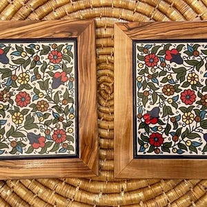 May include: Two square decorative tiles with floral patterns. Each tile features a white background with red, blue, and yellow flowers, green leaves, and brown accents. The tiles are set in wooden frames.