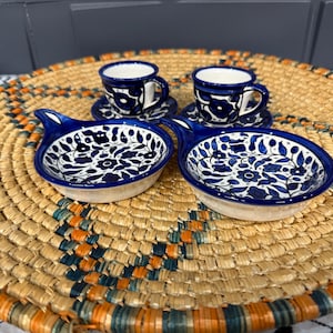 Può includere: Quattro piccole tazze in ceramica blu e bianca con motivi floreali, appoggiate su un sottopentola in paglia intrecciata.