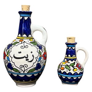 May include: Two ceramic oil cruets with cork stoppers. The larger cruet has a handle and the word "Zayt" in Arabic script. Both are decorated with blue, white, yellow, and red floral patterns. The cruets are likely used for serving olive oil.