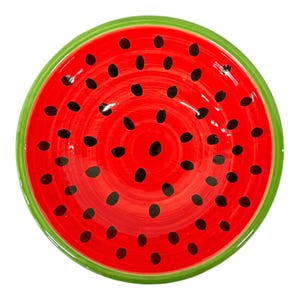 May include: A round ceramic plate with a watermelon design. The plate features a bright red center with black seed details and a green rim. The plate is a decorative item for serving food.