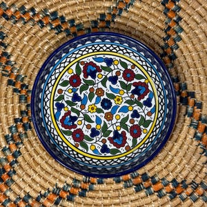 May include: A blue and white ceramic bowl with a floral pattern. The bowl has a white background with blue, red, yellow, and green flowers and leaves. The bowl is surrounded by a woven straw mat.