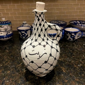 May include: White ceramic bottle with a black and white geometric design and the word "زيت" written in Arabic script. The bottle has a cork stopper and a handle.