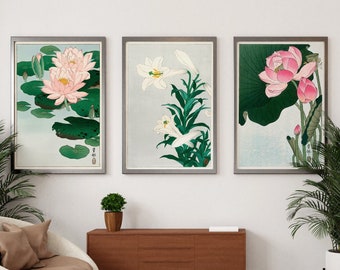 May include: Three framed botanical prints of flowers, including pink water lilies, white lilies, and pink lotus flowers. The prints are hanging on a white wall above a brown wooden cabinet and a white armchair.