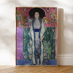 May include: A framed print of a Gustav Klimt painting. The artwork depicts a woman in a long, patterned robe and a large black hat. The background features floral and geometric patterns in shades of green, purple, and pink.