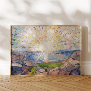 May include: A rectangular framed print featuring an impressionist painting of a bright sunburst over a coastal landscape. The scene displays a central white sun with radiating yellow, orange, and blue light rays above a blue sea and rocky terrain.
