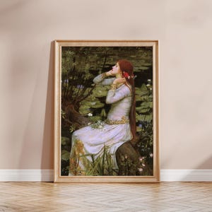 May include: A framed art print depicting a person with long red hair wearing a pale lavender dress with gold trim, seated on a tree trunk near a pond filled with green lily pads and white flowers, set in a light-coloured wooden frame.