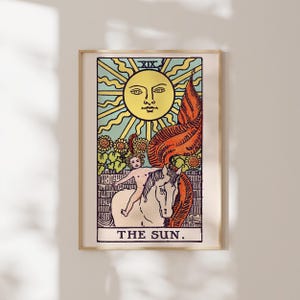 May include: A framed wall art print featuring a tarot card illustration titled THE SUN. The image depicts a large yellow sun with a face above a child riding a white horse, surrounded by sunflowers and a red banner, all set against a light blue sky.