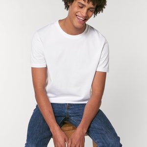 Op de afbeelding: Een wit T-shirt met korte mouwen en een ronde hals. De persoon die het shirt draagt, zit op een houten kruk en draagt een blauwe jeans.