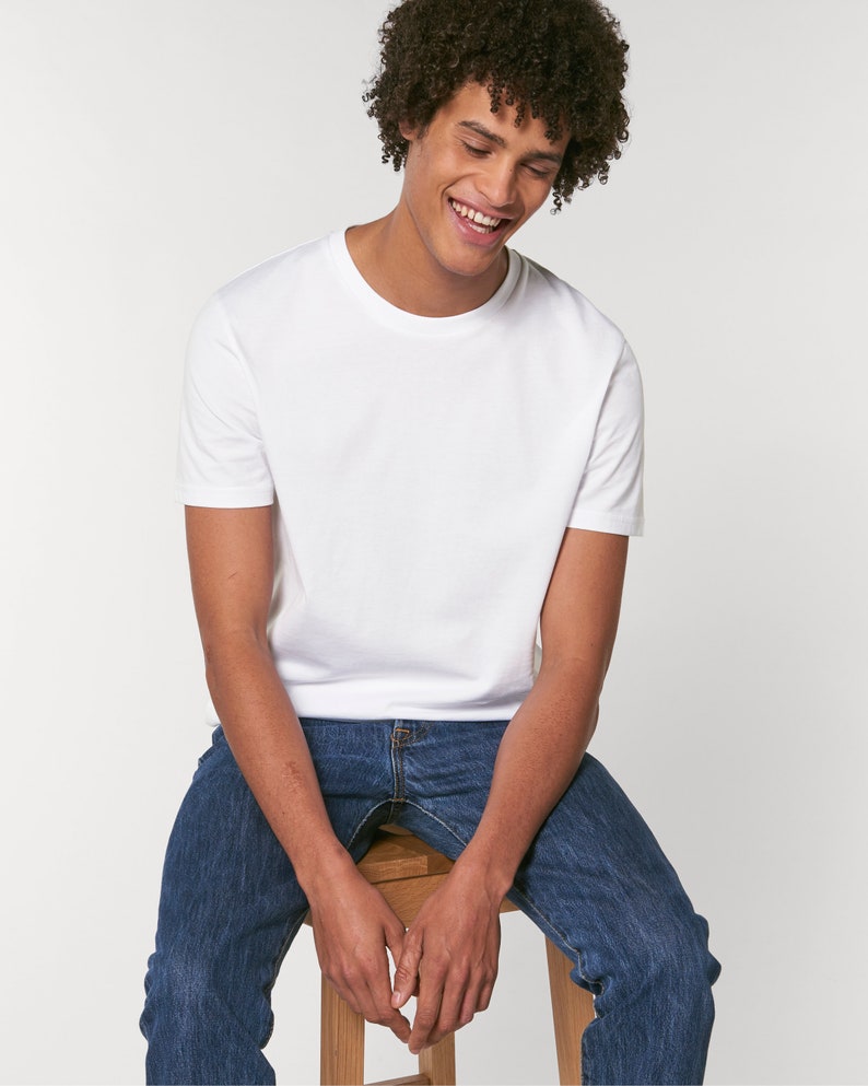 Op de afbeelding: Een wit T-shirt met korte mouwen en een ronde hals. De persoon die het shirt draagt, zit op een houten kruk en draagt een blauwe jeans.