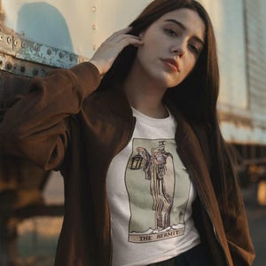 May include: A white t-shirt featuring a graphic print of The Hermit tarot card, which depicts a robed figure holding a lantern, labeled with the Roman numeral IX and the text THE HERMIT, worn under a brown zip-up jacket and black pants.