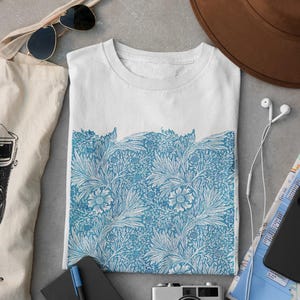 Op de afbeelding: Een wit t-shirt met een blauwe botanische print op de onderste helft, vergezeld van een bruine hoed, zonnebril, canvas tas, zwart notitieboek met blauwe pen, vintage camera, witte bedrade oordopjes en een gevouwen kaart.