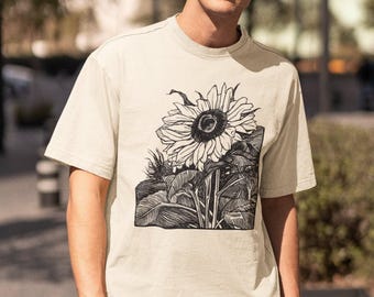 Könnte beinhalten: Ein beiges T-Shirt mit kurzen Ärmeln und einem großen schwarz-weißen Grafikdruck einer Sonnenblume auf der Vorderseite. Das Design zeigt den Blütenkopf mit detaillierten Blütenblättern und Blättern in einem kontrastreichen Stil.
