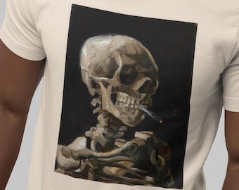 Puede incluir: Una camiseta de color crema con un estampado gráfico rectangular de un cráneo humano con un cigarrillo en la boca. El cráneo está representado en tonos beige, blanco y gris sobre un fondo negro sólido, mostrando una estructura ósea detallada.