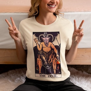 May include: A cream-colored short-sleeve t-shirt featuring a graphic print of The Devil tarot card. The design displays a horned figure with wings, a pentagram above its head, and two smaller figures chained to a pedestal, with the text THE DEVIL.