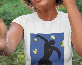 Może przedstawiać: Biały t-shirt z krótkim rękawem z prostokątnym nadrukiem graficznym na klatce piersiowej. Nadruk przedstawia stylizowaną czarną ludzką sylwetkę na niebieskim tle, ozdobioną kilkoma żółtymi kształtami gwiazd i małą czerwoną kropką na klatce piersiowej.