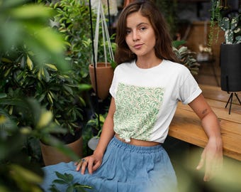 Może przedstawiać: Biały t-shirt z krótkim rękawem z prostokątnym panelem z przodu, ozdobionym botanicznym wzorem liści w kolorach zielonym i kremowym. Koszulka jest zawiązana w talii i zestawiona z długą, zwiewną niebieską spódnicą z tkaniny o wyraźnej fakturze.