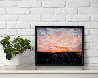 May include: A framed landscape painting featuring a vibrant sunset with pink, orange, and blue clouds above a dark, silhouetted cityscape. The artwork is displayed in a simple black frame, positioned next to a green plant in a white pot.