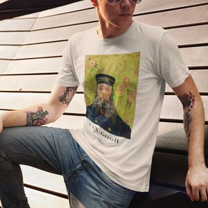 Op de afbeelding: Een wit T-shirt met een print van een schilderij van Vincent van Gogh. Het schilderij toont een man met een baard die een blauw uniform en een pet draagt. De achtergrond is een geel en groen bloemenpatroon. De tekst "VAN GOGH" is onder het schilderij gedrukt.
