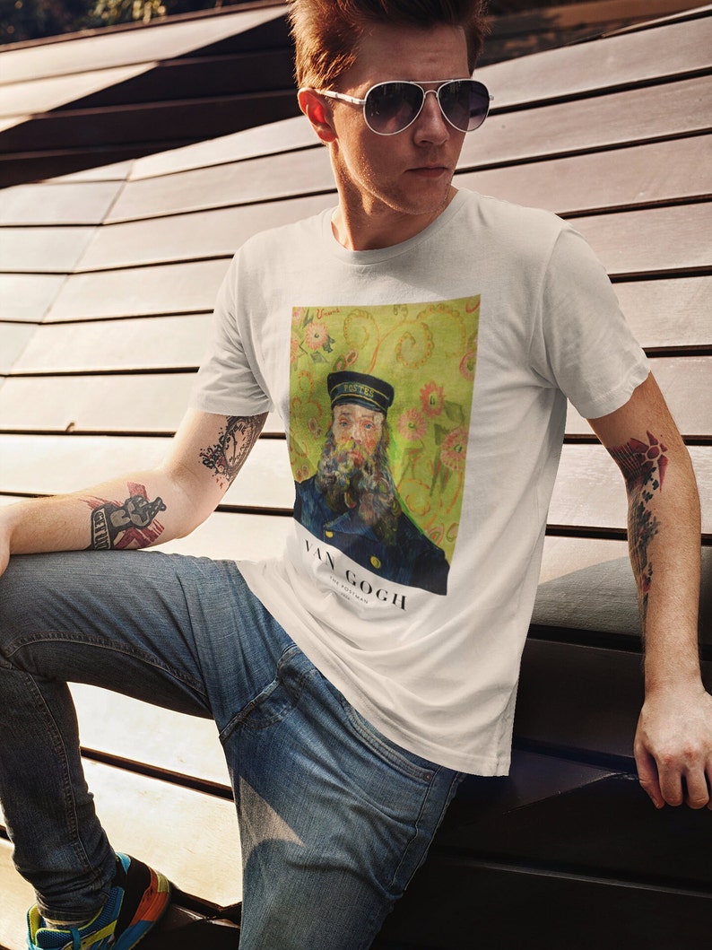 Op de afbeelding: Een wit T-shirt met een print van een schilderij van Vincent van Gogh. Het schilderij toont een man met een baard die een blauw uniform en een pet draagt. De achtergrond is een geel en groen bloemenpatroon. De tekst "VAN GOGH" is onder het schilderij gedrukt.