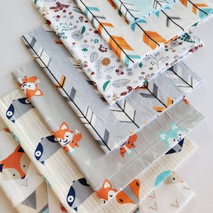 May include: A set of eight fabric squares with various patterns, including arrows, flowers, and woodland animals. The fabrics are in shades of gray, orange, blue, and white.
