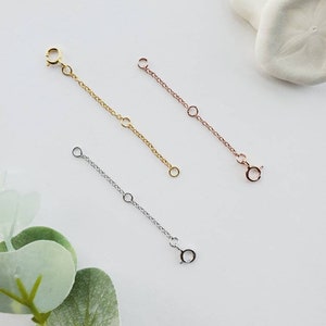 May include: A selection of necklace extenders in gold, rose gold, and silver. Each extender has a small chain with a clasp at one end and a loop at the other, designed to lengthen necklaces. The extenders are displayed on a white surface.