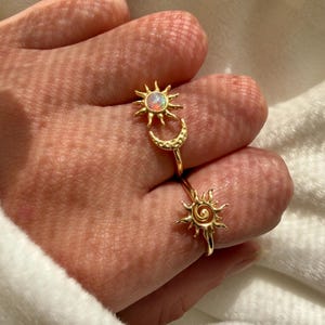 May include: Two gold-toned rings worn on fingers. One ring features a sun motif with a central iridescent opal-like stone paired with a crescent moon. The second ring displays a sun design with a spiral center and radiating pointed rays.