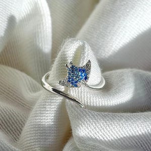 May include: A silver-toned open ring featuring a small, triangular-shaped charm encrusted with tiny, sparkling blue crystals. The ring has a slender, polished band that curves around to form an adjustable, bypass-style design with a metallic finish.