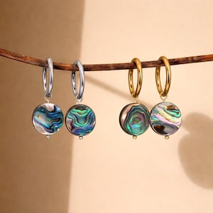 Op de afbeelding: Twee paar oorringen met iriserende abalone schelpen hangers. Eén paar is zilverkleurig, de andere goudkleurig. De abalonschelpen tonen een mix van blauwe, groene en paarse tinten, hangend aan de oorringen.