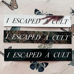 May include: Three rectangular bumper stickers featuring the text I ESCAPED A CULT in a white serif font. The top sticker is white with a dark smudge, the middle has a dark textured pattern, and the bottom sticker is solid black.