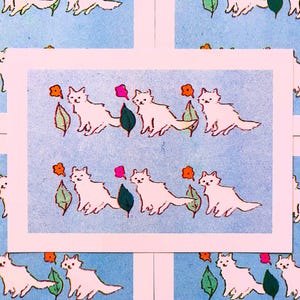 May include: A rectangular print featuring a repeating pattern of white cats on a light blue background. The design includes small orange flowers and green leaves interspersed between the cats, arranged in two horizontal rows across the paper.