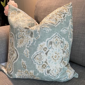 May include: A decorative throw pillow with a floral pattern in shades of blue, beige, and brown. The pillow is placed on a gray sofa, showcasing its design and texture. The pillow's pattern features stylized floral and leaf motifs.