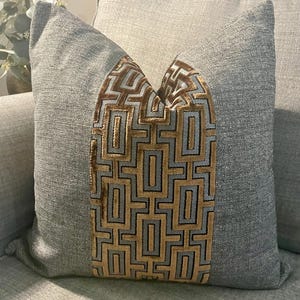 May include: A grey cushion with a geometric gold and black pattern. The cushion is made of textured fabric and is designed for home decor. The rectangular cushion is placed on a grey sofa.