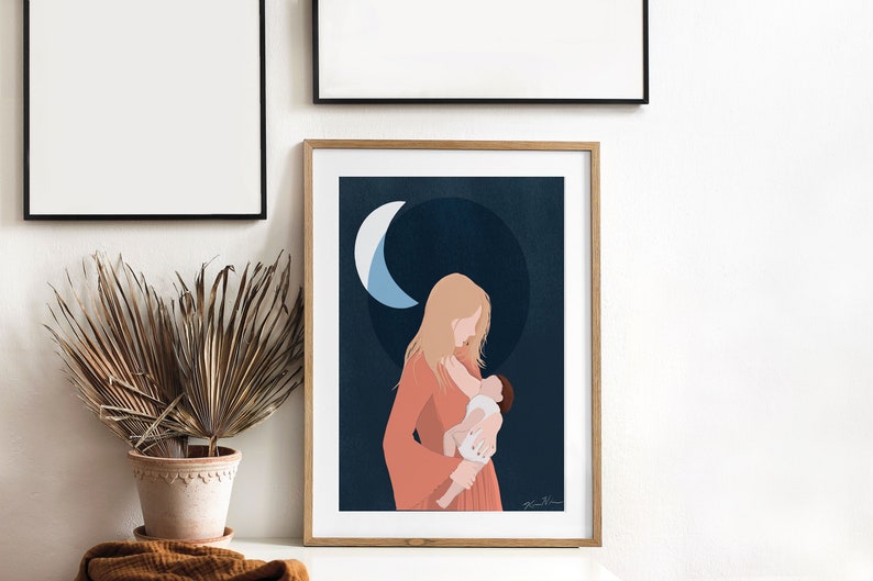 Op de afbeelding: Een ingelijste kunstafdruk toont een moeder die een baby vasthoudt, tegen een donkerblauwe achtergrond met een halve maan. De moeder draagt een koraalkleurige jurk. De print zit in een houten lijst.