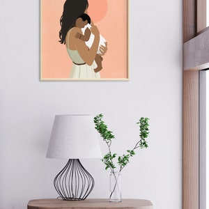 Op de afbeelding: Een ingelijste kunstdruk toont een moeder die een baby vasthoudt. De illustratie toont een vrouw met donker haar en een lichtgroene jurk, die een baby in een witte onesie vasthoudt. De achtergrond is perzik met twee witte en perzik cirkels.