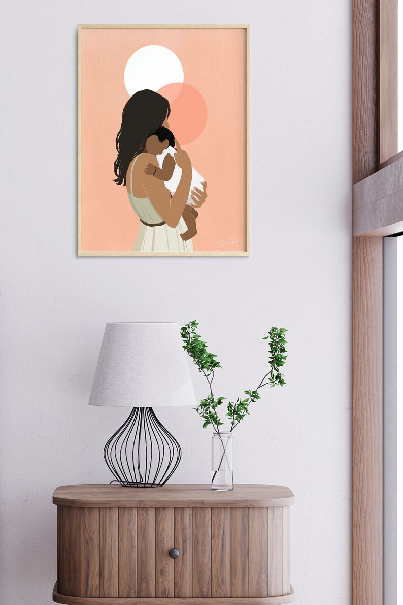 Op de afbeelding: Een ingelijste kunstdruk toont een moeder die een baby vasthoudt. De illustratie toont een vrouw met donker haar en een lichtgroene jurk, die een baby in een witte onesie vasthoudt. De achtergrond is perzik met twee witte en perzik cirkels.
