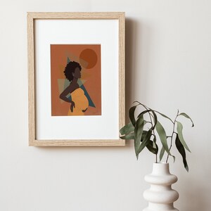 Op de afbeelding: Ingelijste kunstdruk met een silhouet van een zwangere persoon in een gele jurk, tegen een oranje achtergrond met geometrische vormen en een zon. Het kunstwerk bevindt zich in een licht houten lijst, tentoongesteld aan een witte muur. Een witte vaas met groene bladeren staat op een houten tafel.
